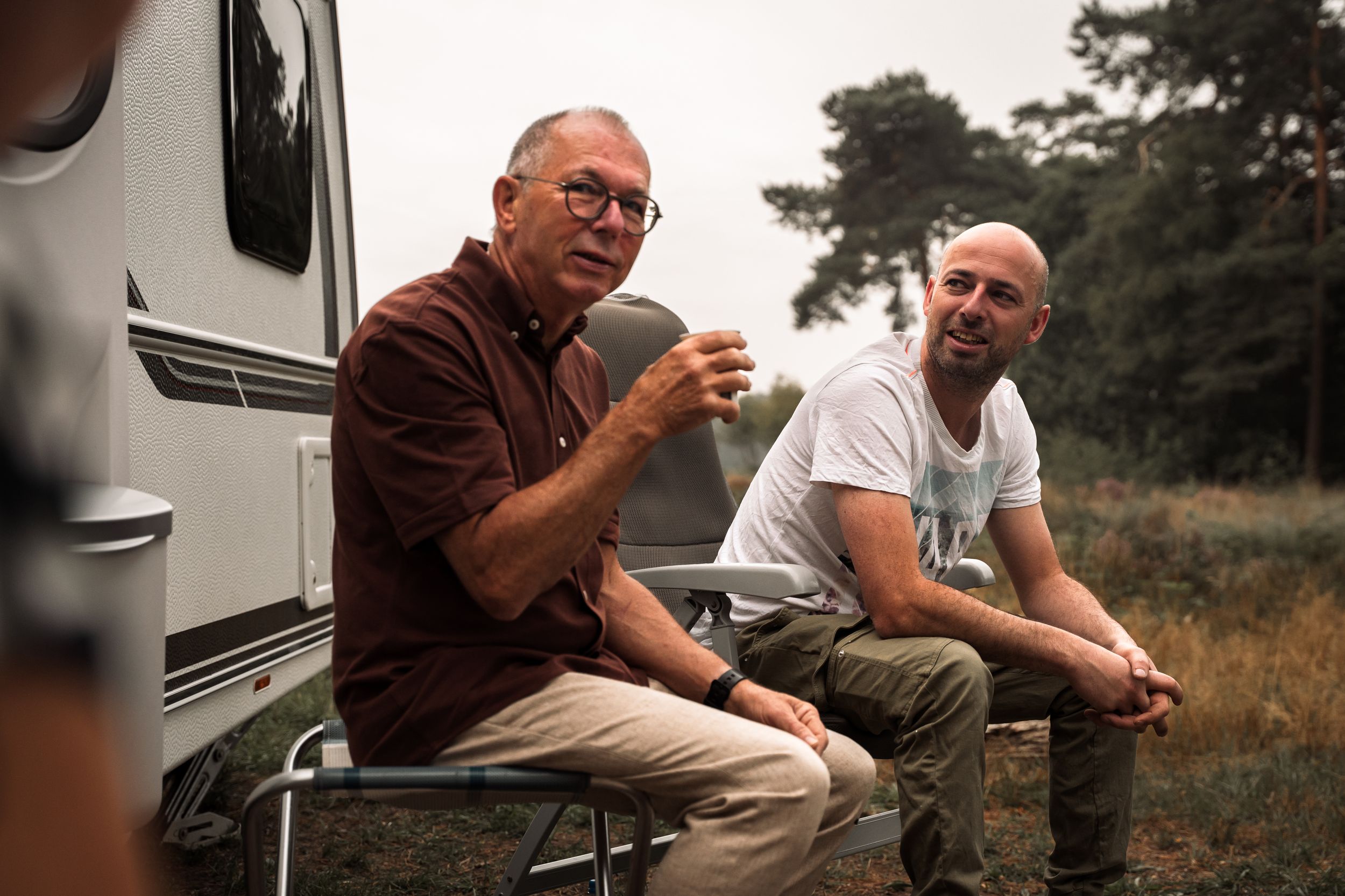 Two people sit and chat outside an RV camper in a natural outdoor setting during golden hour.