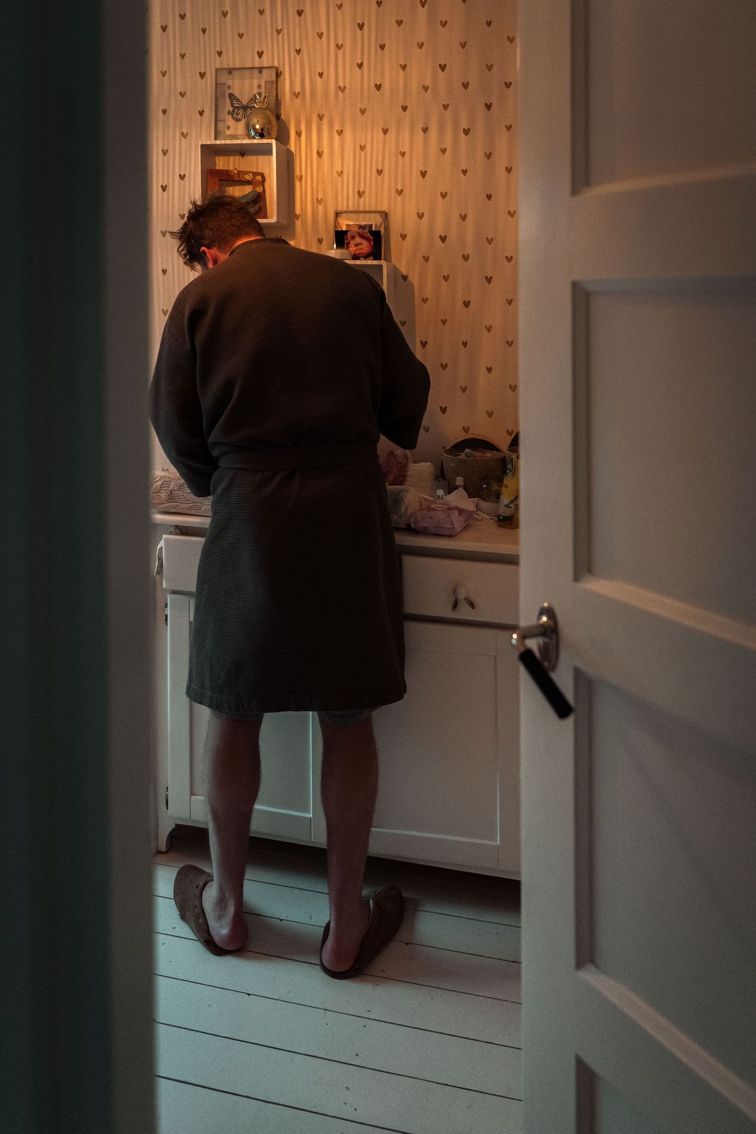 A person in dark clothing bends down to look inside a lower cabinet in a room with striped wallpaper.