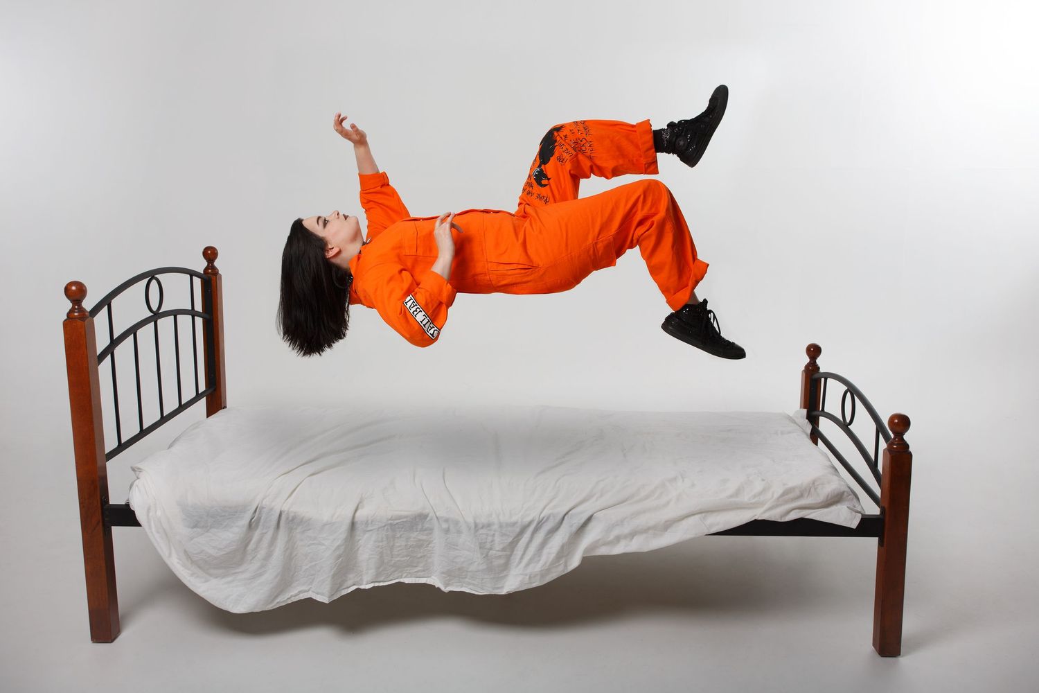 A person in bright orange jumpsuit levitates horizontally above a bed with white sheets and wooden posts.