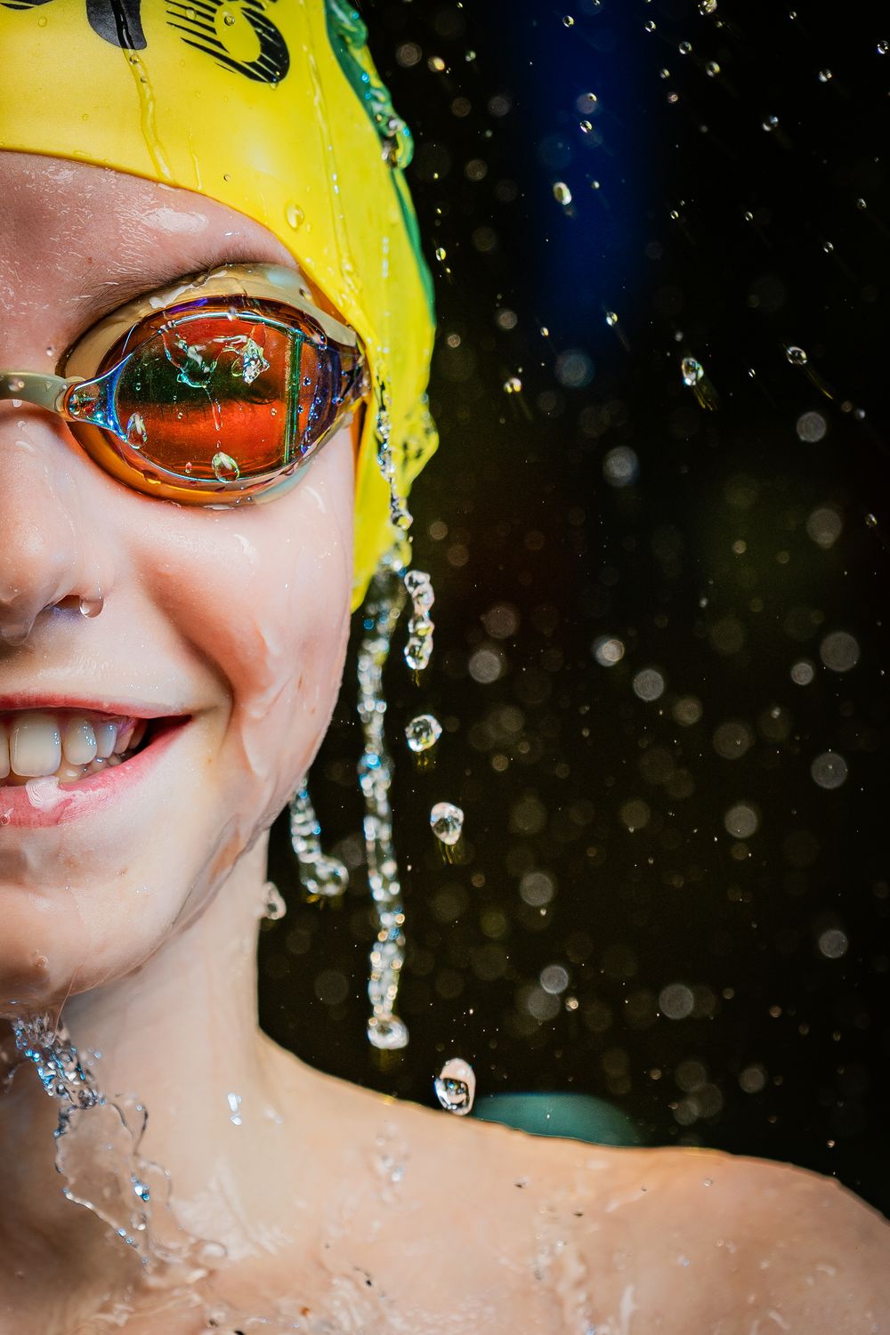 Smiling young swimmer in yellow cap and orange goggles with water splashing around.