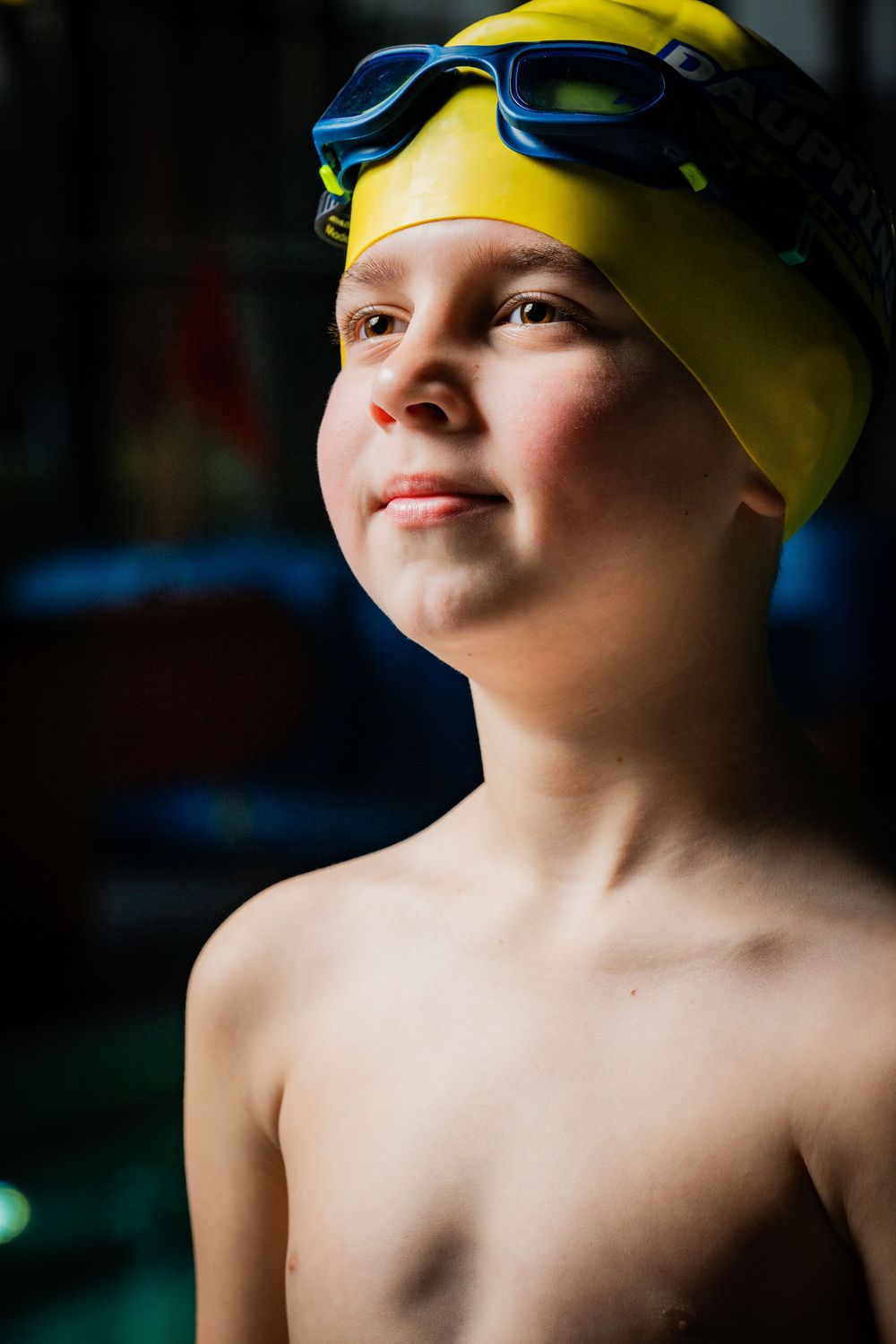 A young boy wearing a yellow swim cap and blue goggles smiles confidently at an indoor pool.