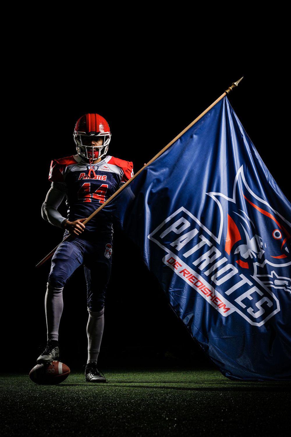 American football player holding a Patriots de Ribeauville team flag on dark field.