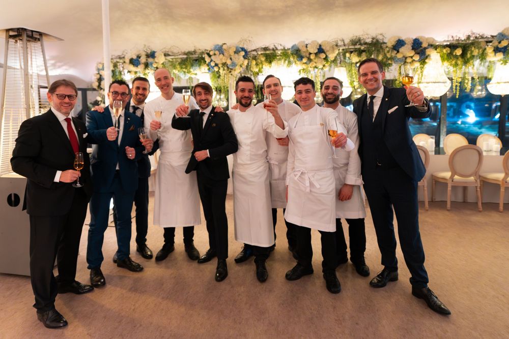A group of chefs and staff members pose together at an elegant event venue with floral decorations and lighting.
