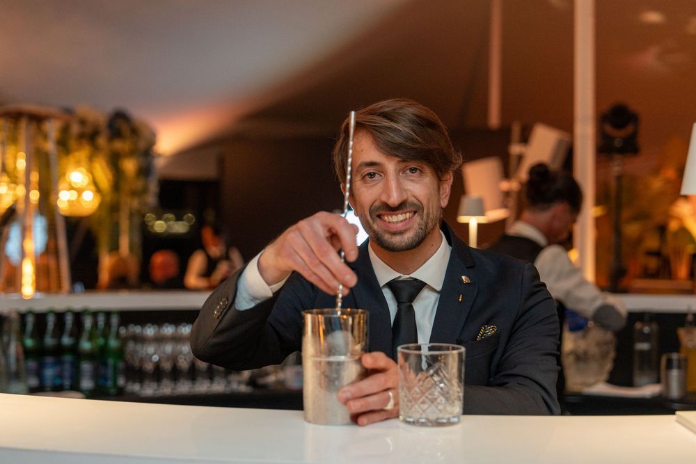 Professional bartender in black suit mixing a cocktail with precision at an elegant bar counter.