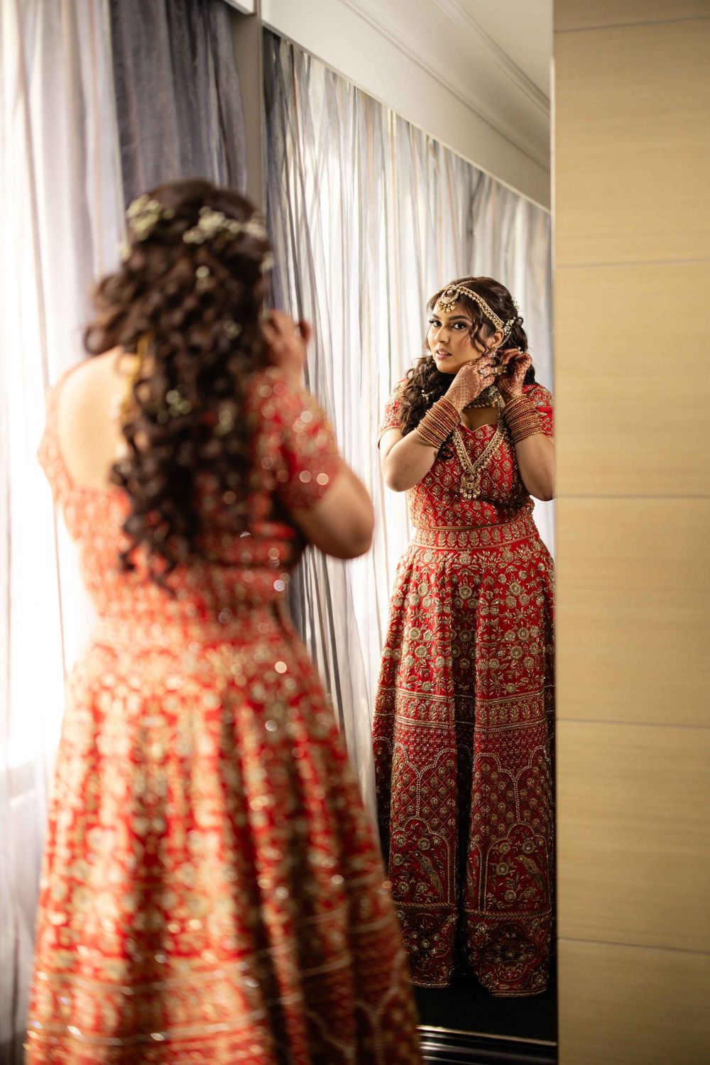 Indian bride in red embroidered lehenga adjusts earring while looking at her reflection in mirror.