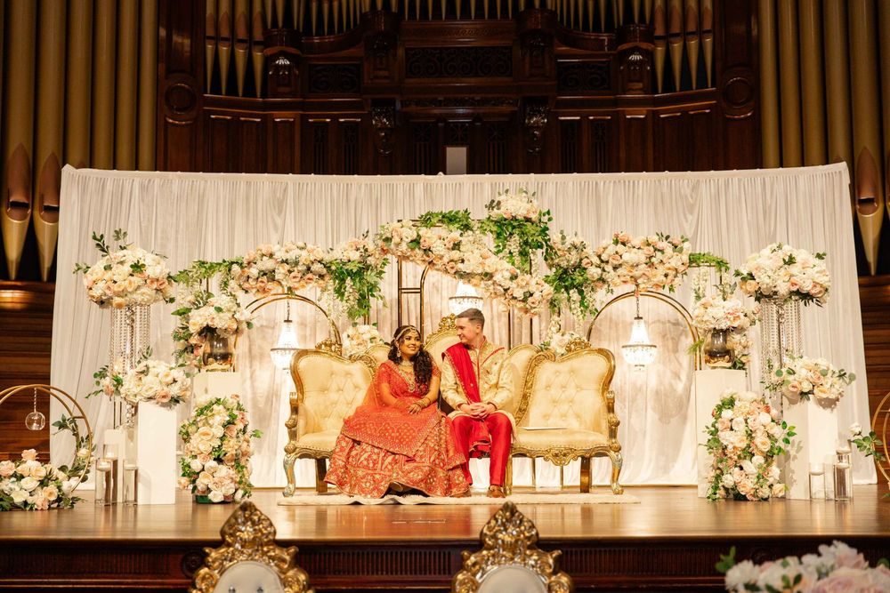Indian bride and groom sit on golden thrones before white floral backdrop in grand wedding hall.