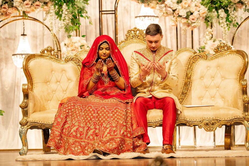 Indian bride and groom in traditional red and gold attire pray together on golden throne chairs.