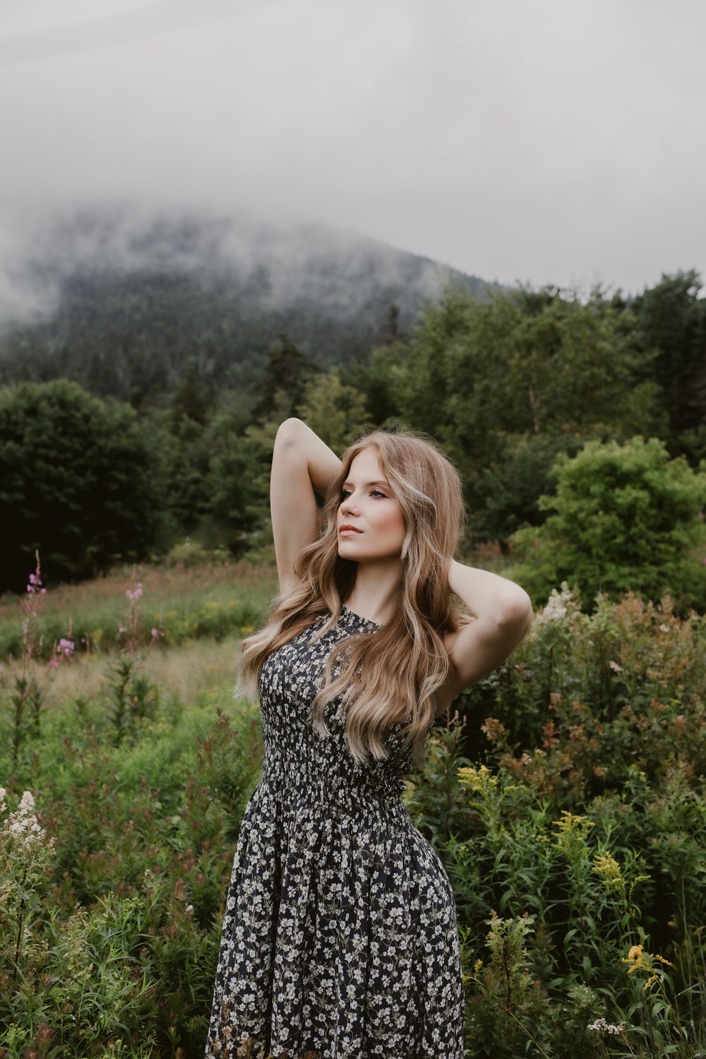 Rachel Delong, Christian musician, wearing a gray floral dress, poses in a field on a foggy day with a hill in background in Lincoln, NH.