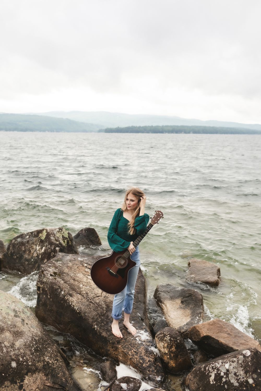 Rachel Delong, Christian musician, stands on a rock holding a guitar at Newfound Lake in Hebron, New Hampshire for portraits.