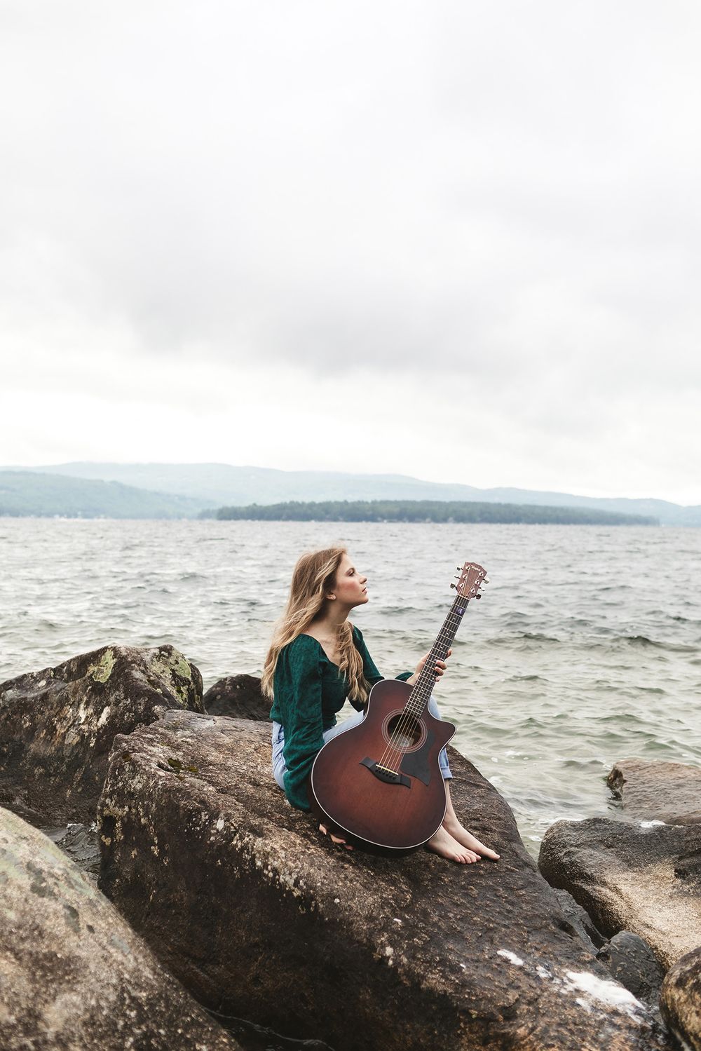 Rachel Delong, Christian musician, poses on a rock sitting for a portrait holding her guitar at Newfound Lake, Hebron New Hampshire.