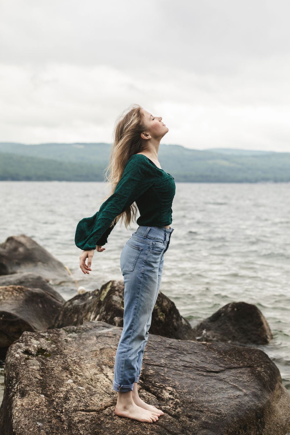 Rachel Delong, Christian musician, wears an emerald green shirt and blue jeans and poses gracefully standing on a rock at Newfound Lake NH.