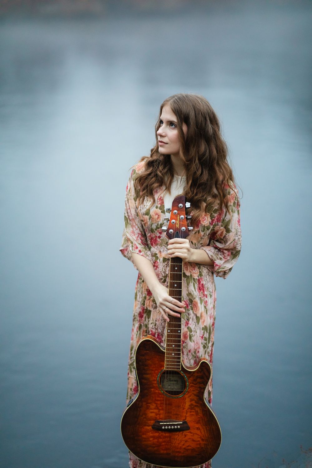 Rachel Delong, Christian musician, wears a floral dress holding her guitar next to a blue colored river in Concord, Massachusetts.