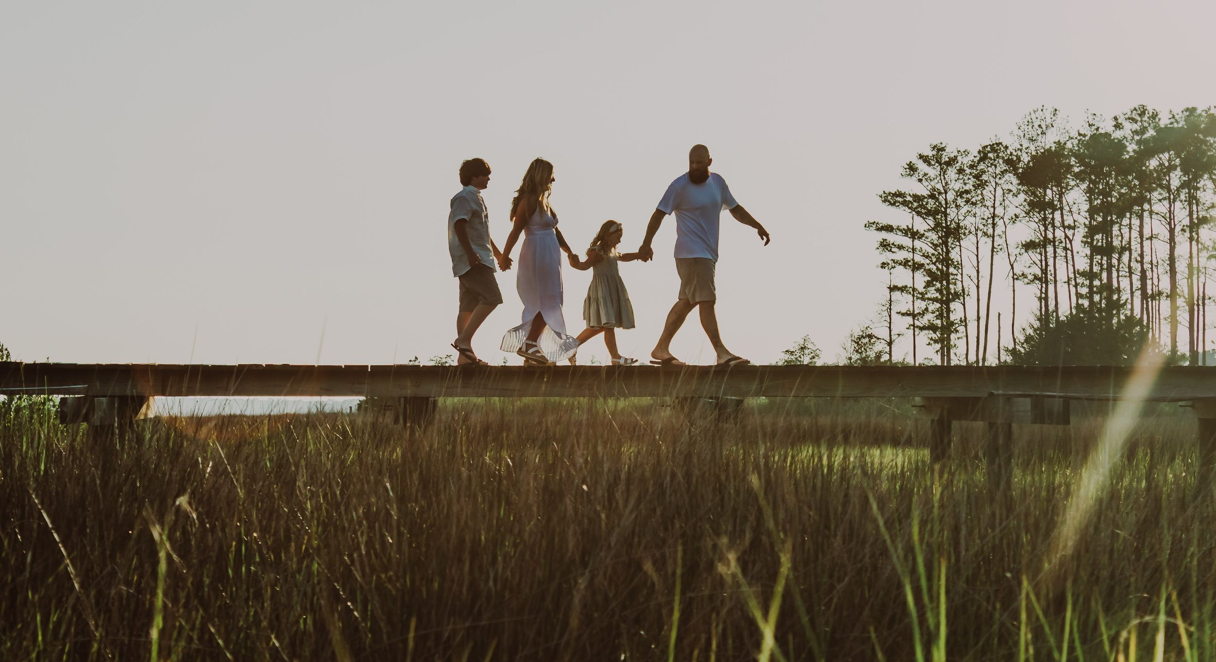 family on a pier holding hands with the sun in the background and the river grass showing