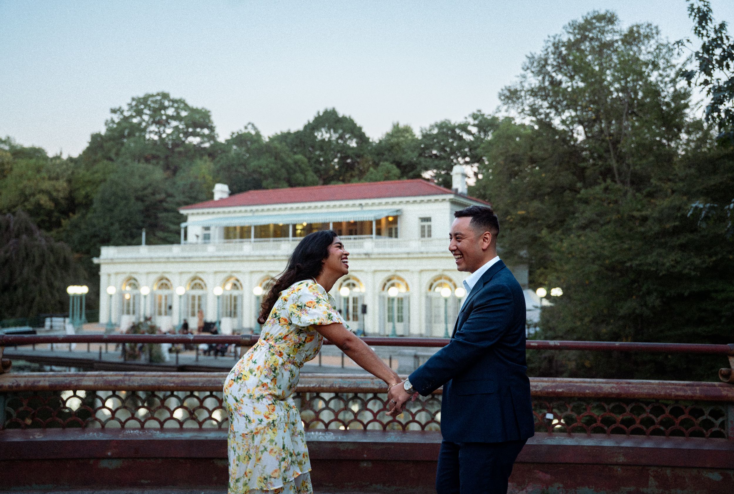 A romantic couple poses together on a bridge with a grand white mansion visible in the background at dusk.