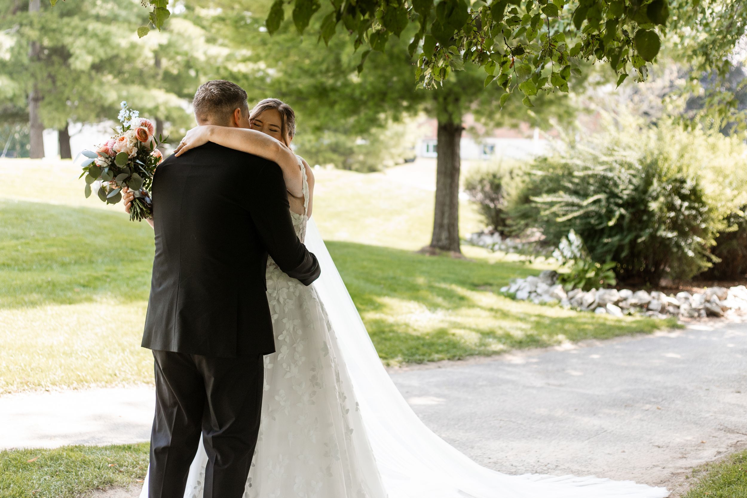 A couple embraces on their wedding day in a sunlit garden setting with a flowing white bridal gown and black tuxedo.