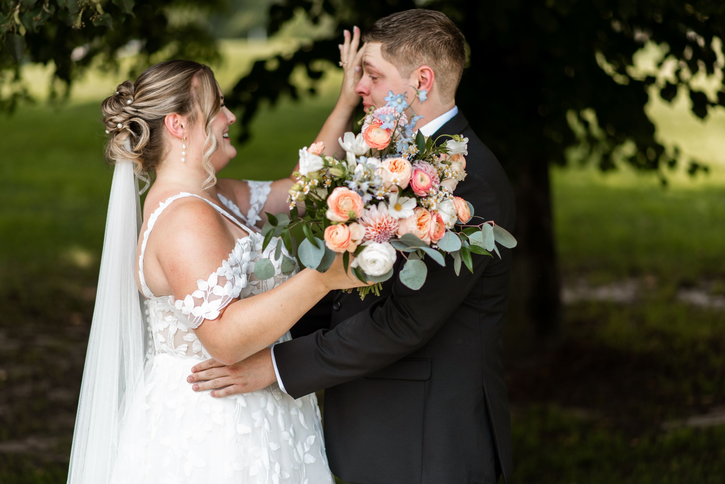 A romantic outdoor wedding scene with a couple embracing under lush green trees while holding a pink and white bouquet.