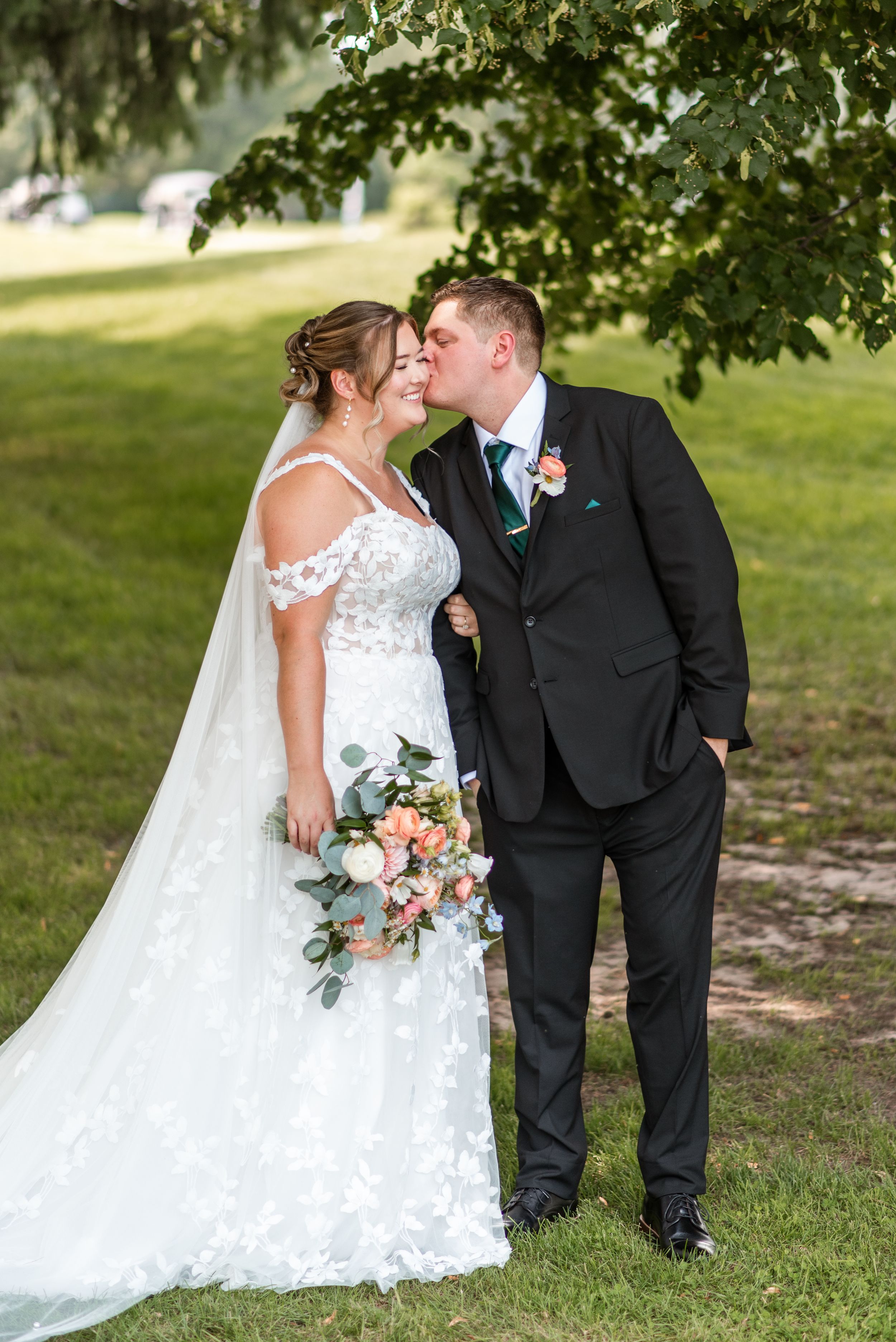A romantic wedding couple shares intimate kisses under green trees in a park setting.