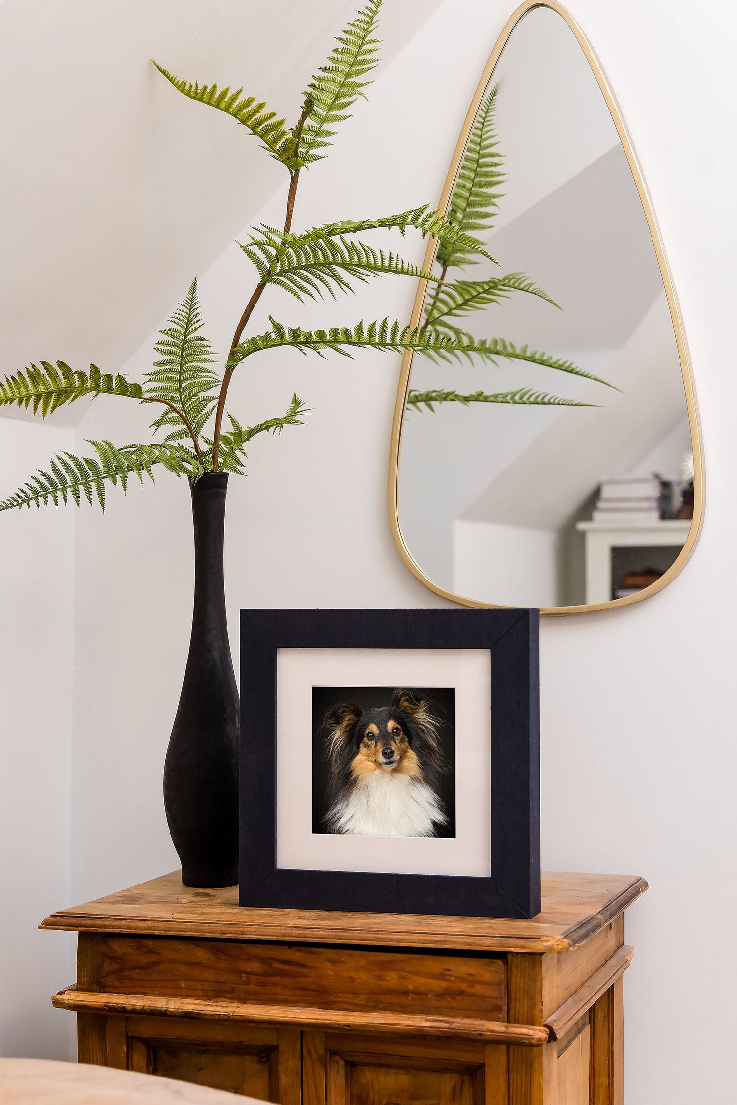 Modern home decor vignette with black vase holding ferns, framed photo, and triangular mirror on wooden sideboard.