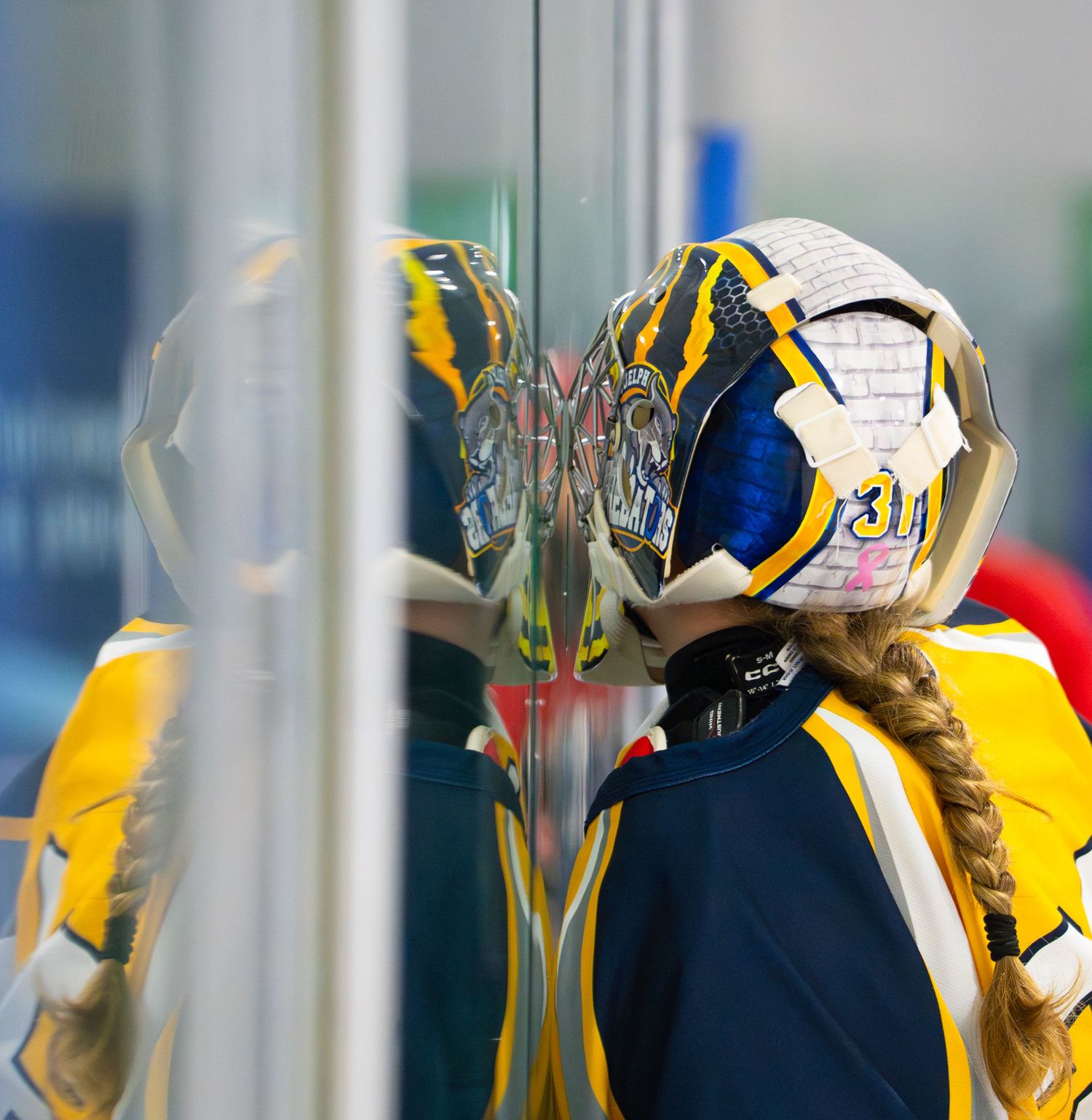 Reflection of ringette player in blue and yellow uniform seen in glass barrier.