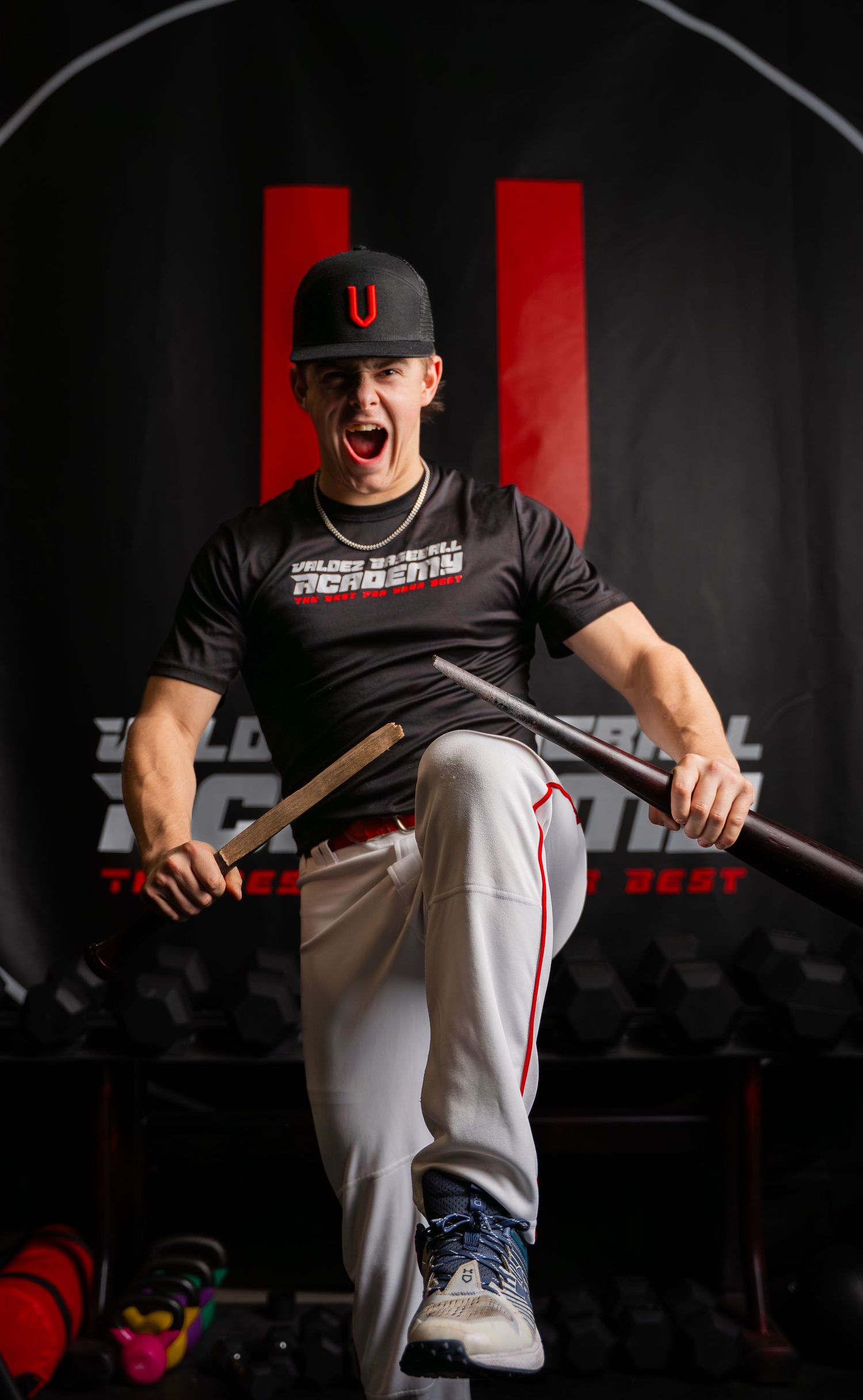 Baseball player in black uniform and cap shows intense expression while holding a bat in dramatic lighting.