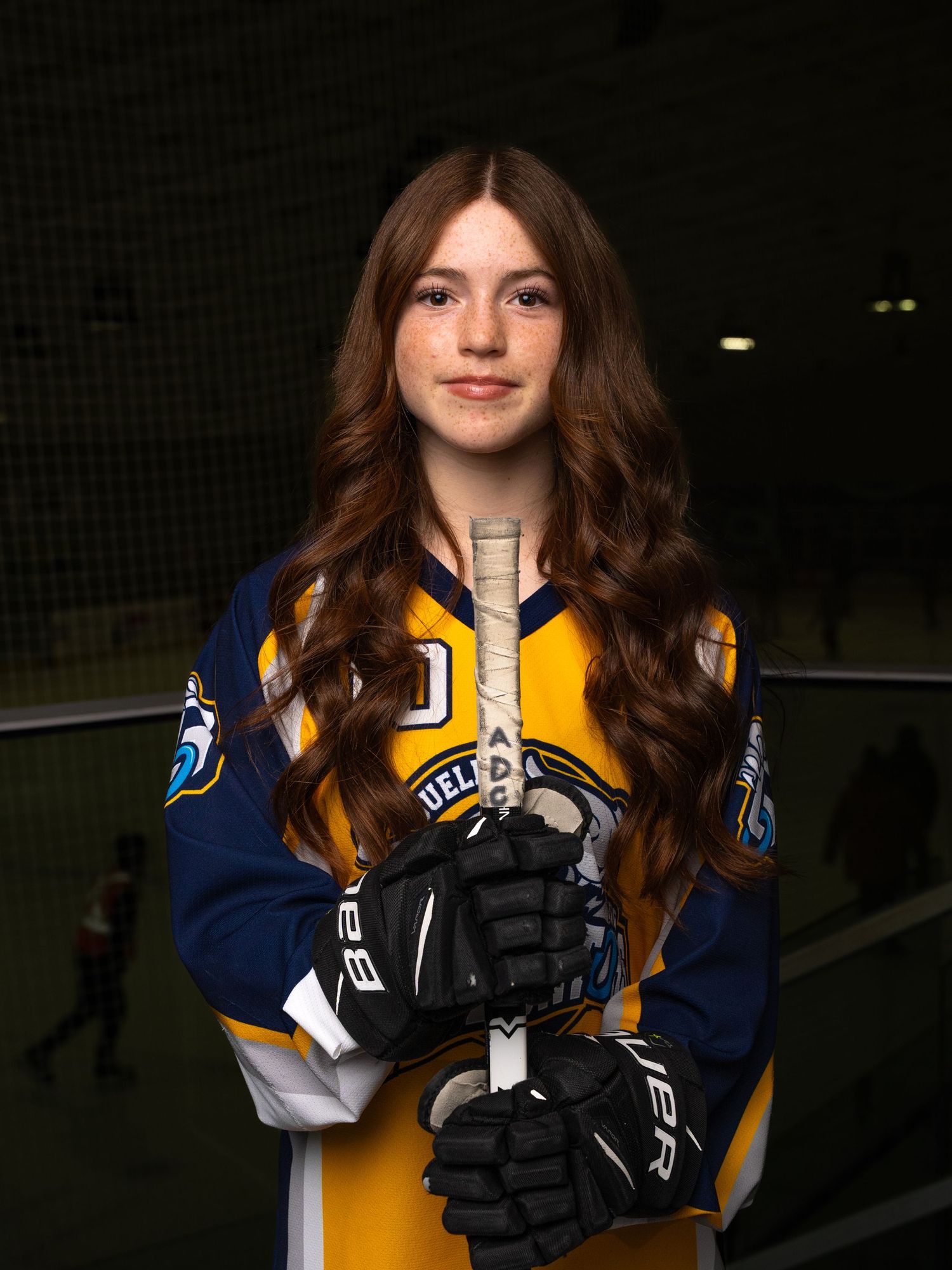 Ringette player in blue and yellow jersey with long hair poses for portrait in dark setting.