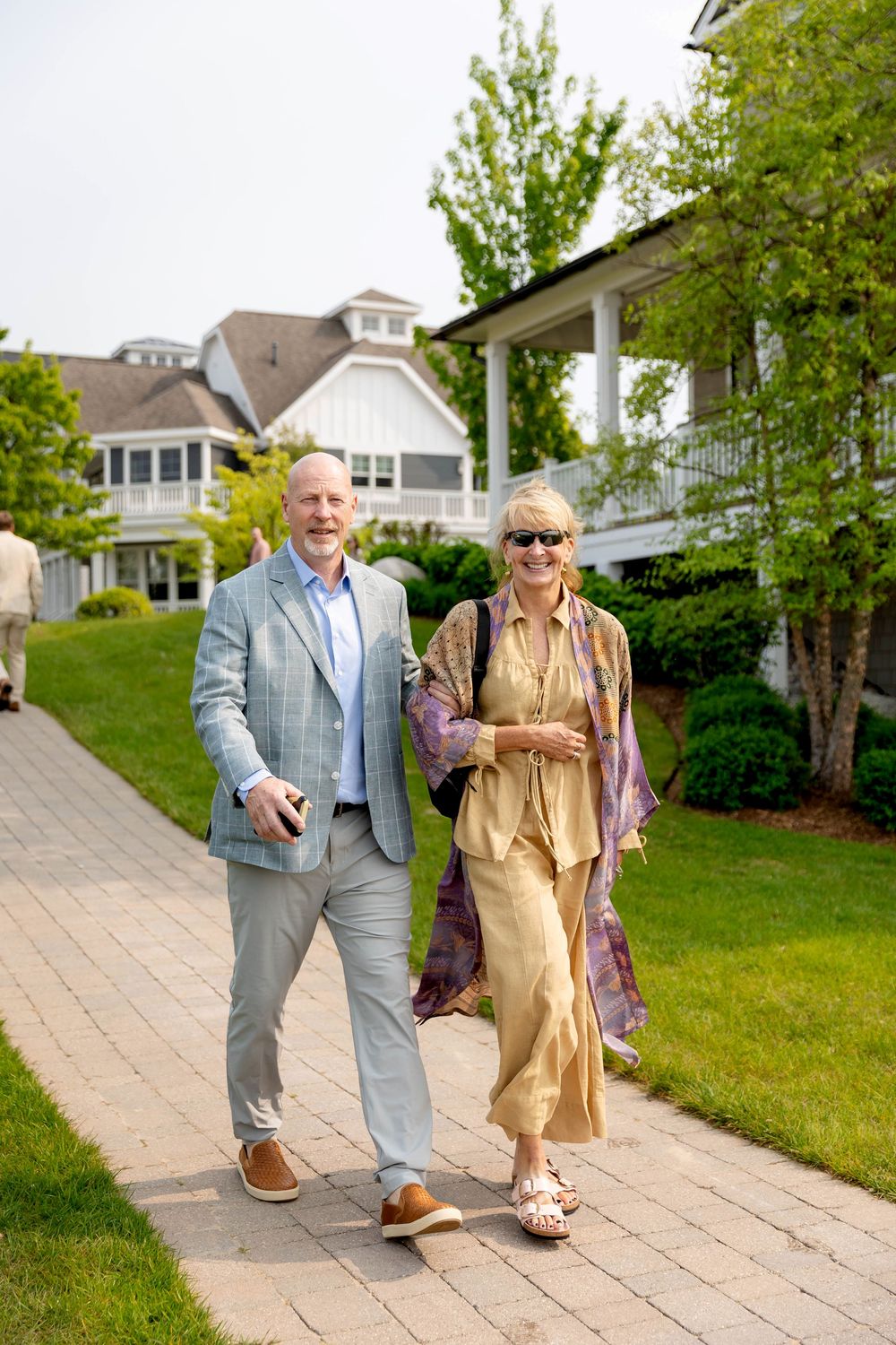 Two seniors walk together on a paved garden path surrounded by green grass and trees on a sunny day.