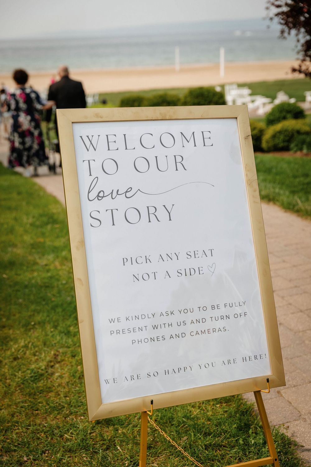 A wedding welcome sign on an easel reads 'Welcome to our love story' with beach and ocean views in the background.
