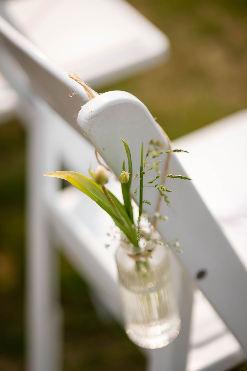 Small glass vase with white calla lily and baby's breath flowers hangs on white wooden chair at outdoor event.