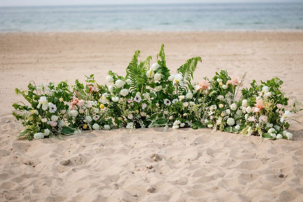 A lush floral arrangement with white flowers and greenery decorates the sandy beach with ocean waves in the background.