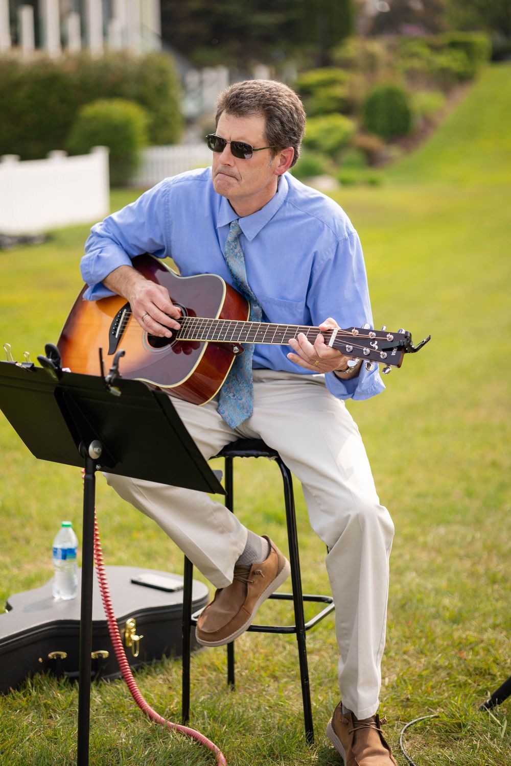 Musician in blue shirt and white pants plays acoustic guitar while seated outdoors on folding chair in grassy setting.