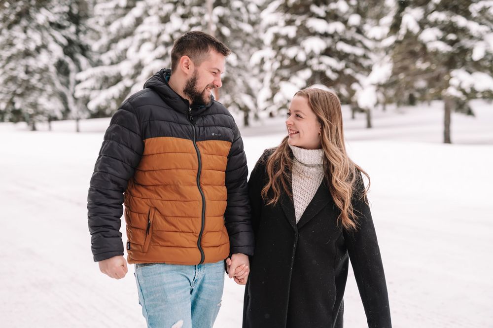 A couple in winter coats walks together through a snowy landscape while smiling and holding hands.