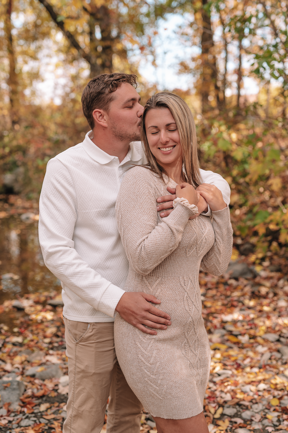 A romantic autumn engagement photo shows a couple embracing among fallen leaves in a wooded setting.