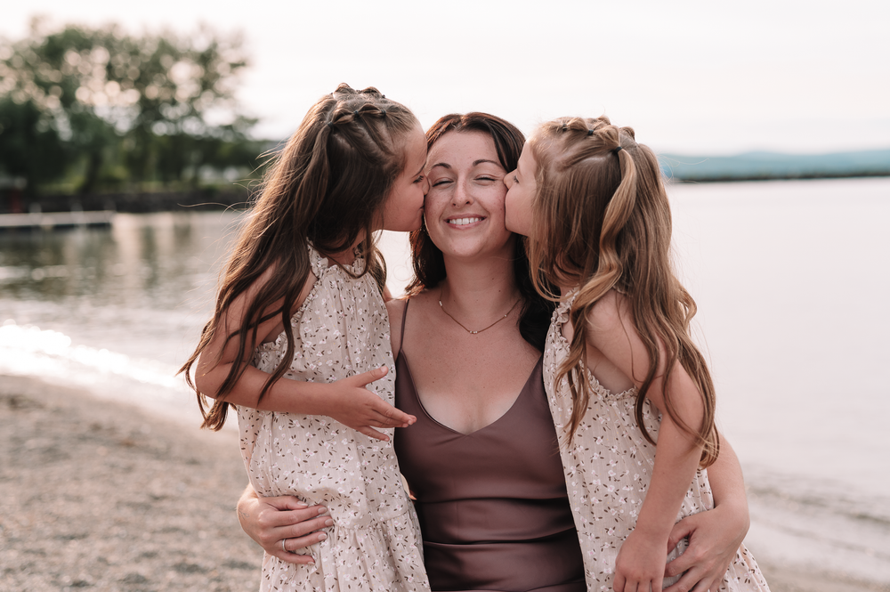 Three people share a tender moment on a beach at sunset with a lake in the background.