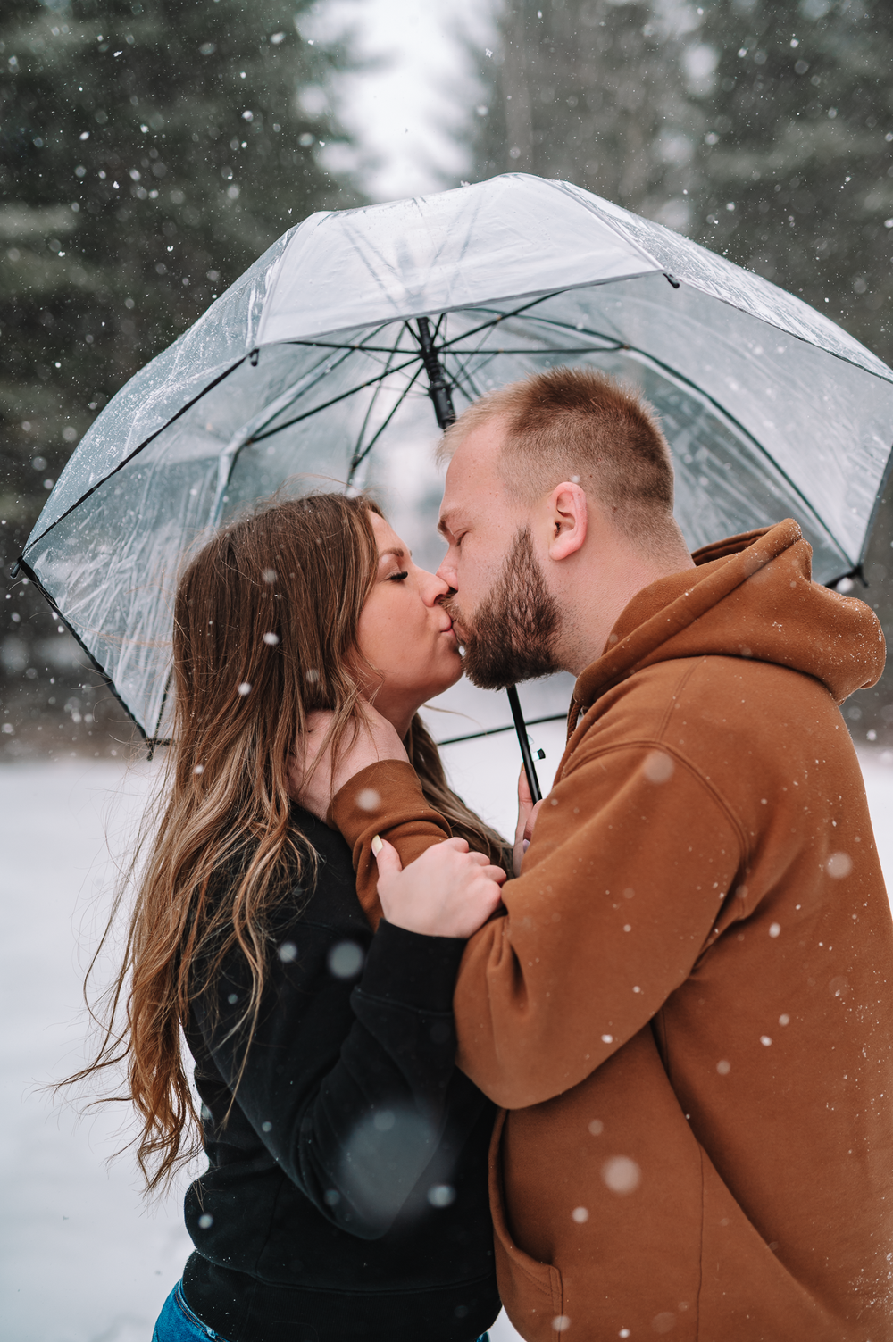 A romantic couple shares an intimate moment under a clear umbrella while snow falls gently around them in winter.