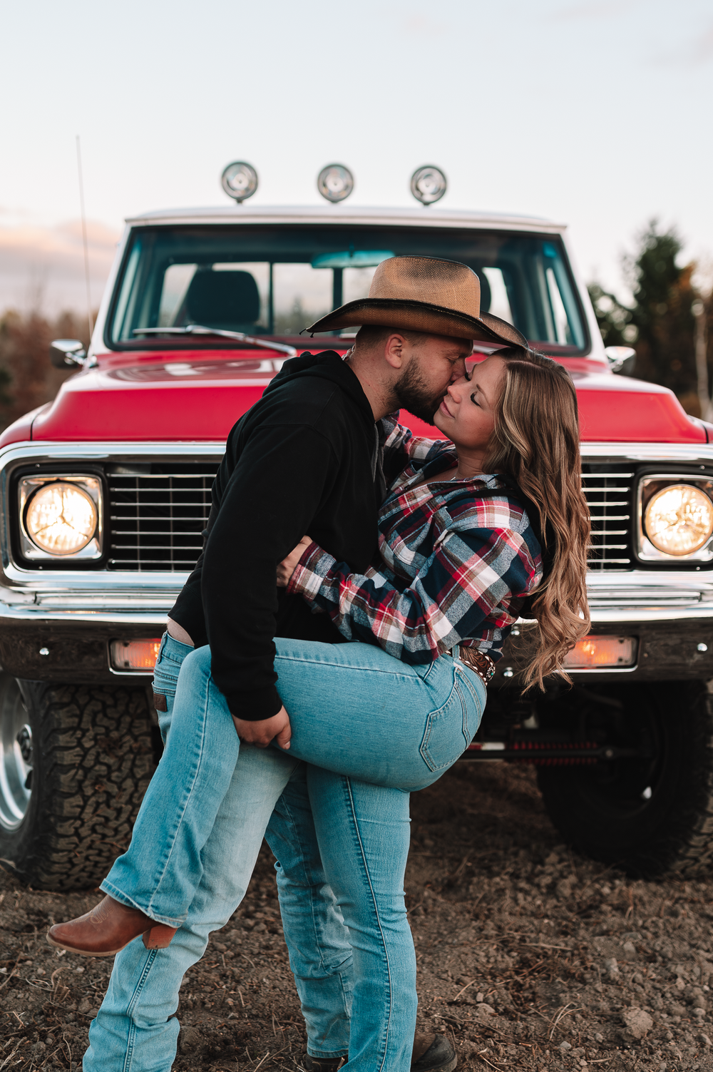 A romantic couple embraces in front of a vintage red pickup truck with off-road tires at sunset.