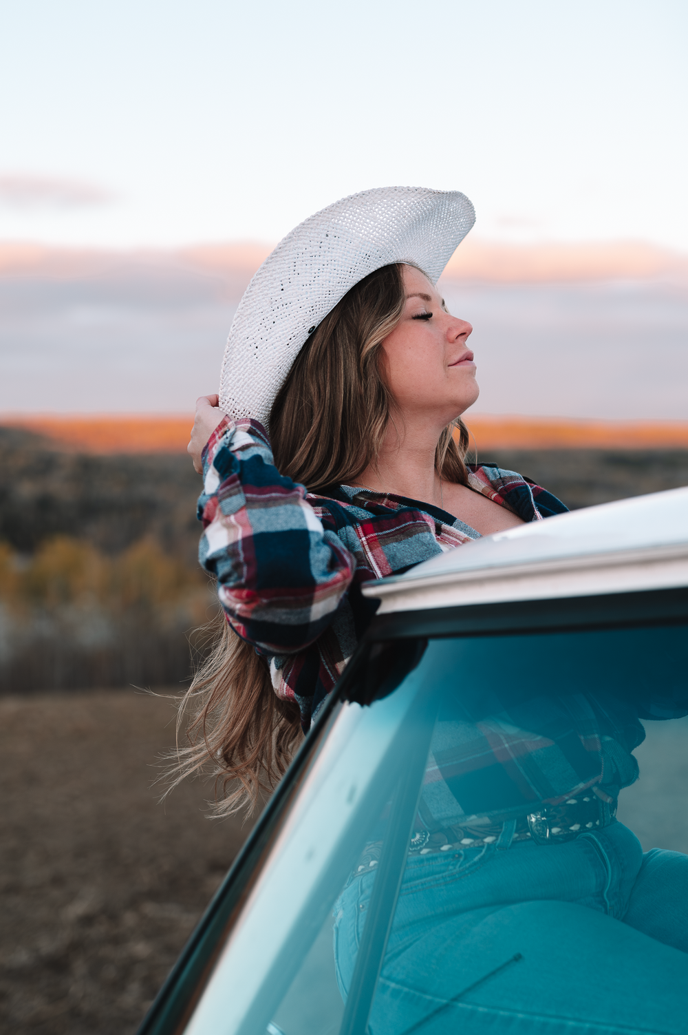 Person with long hair wearing a plaid winter hat leans back against a turquoise car during sunset.