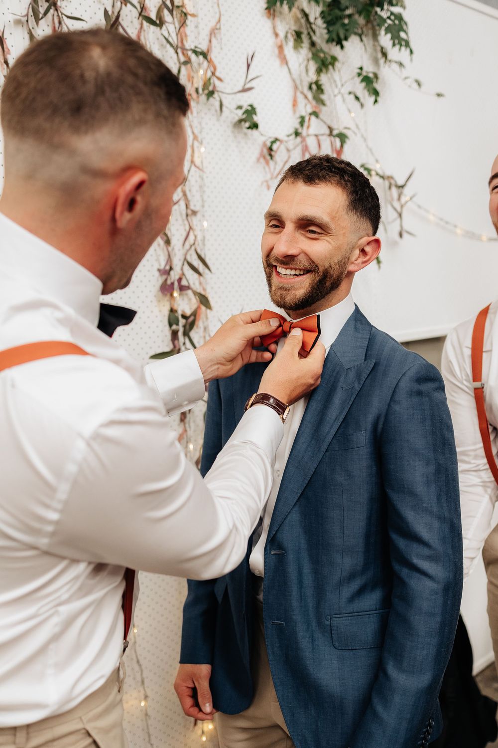 Wedding party members share candid moments and laughter against white wall with greenery decorations.