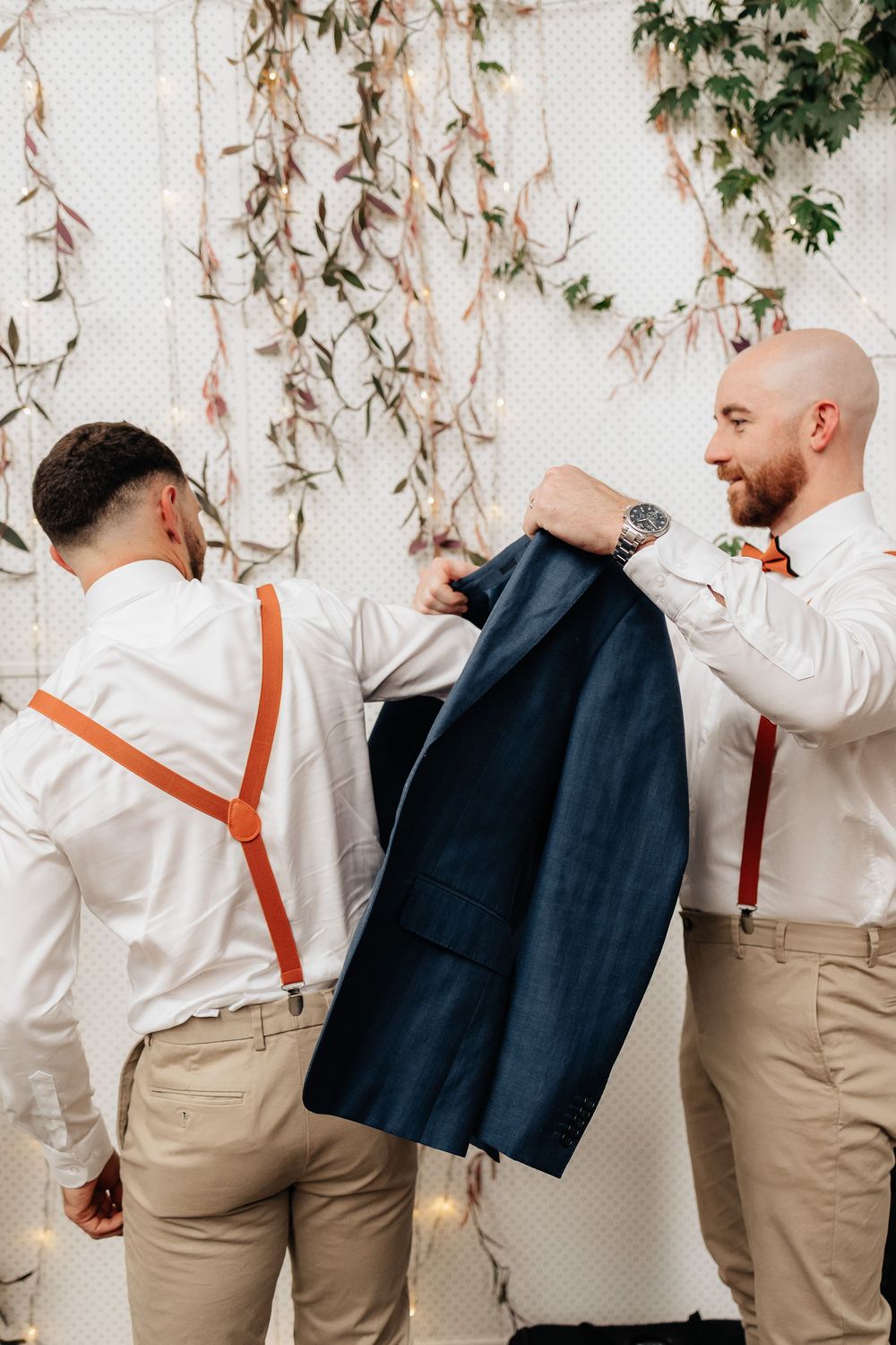 Groomsmen in khaki pants and white shirts with orange suspenders posing against a white backdrop with hanging greenery.