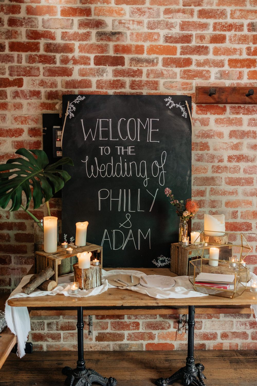 Welcome chalkboard signs and candle decorations against rustic brick wall at wedding reception.