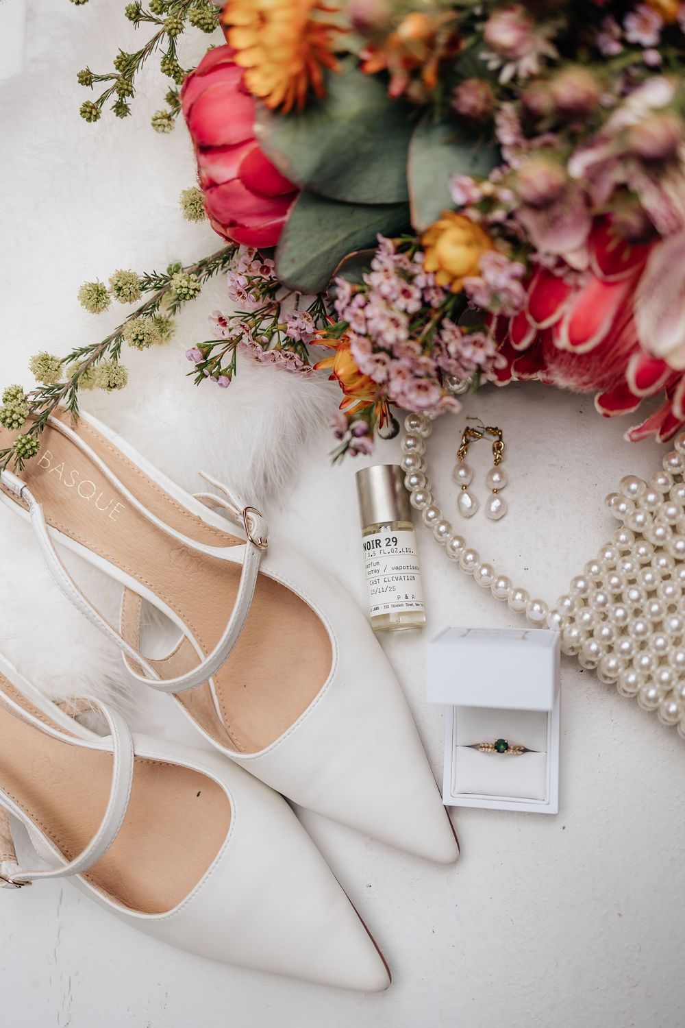 Wedding accessories including white heels, flowers, pearls and rings arranged on white surface.