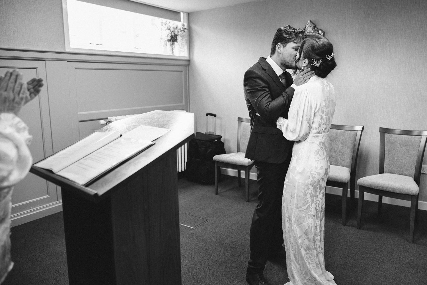 A couple shares a romantic kiss during their wedding ceremony in a small chapel room with a podium and chairs.