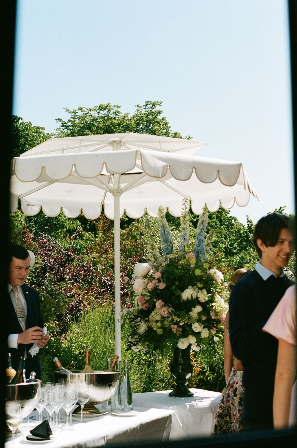 Outdoor catering setup with white umbrellas and floral arrangements on buffet tables.