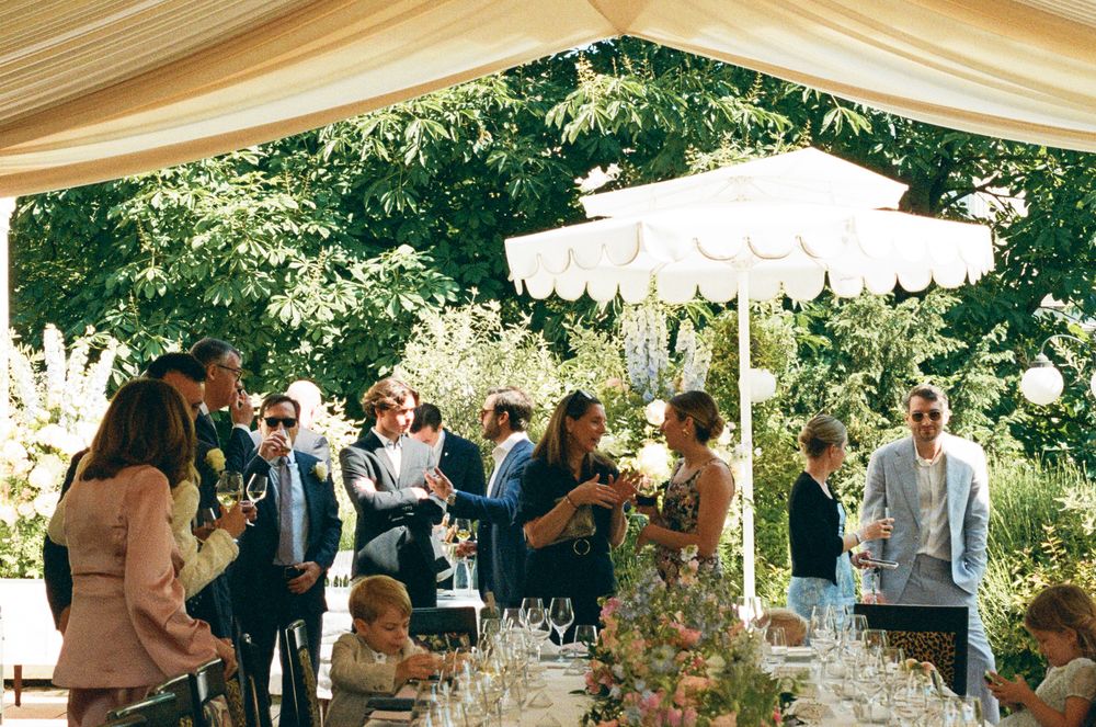 A panoramic view of an outdoor wedding reception with white tents, tables, and guests mingling in a garden setting.