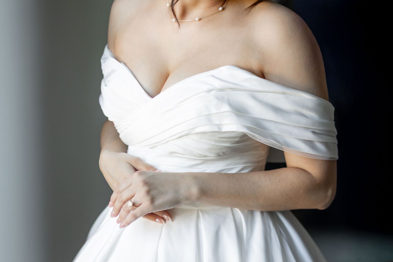 Close-up of an elegant white off-shoulder wedding dress with ruched bodice detail against dark background.