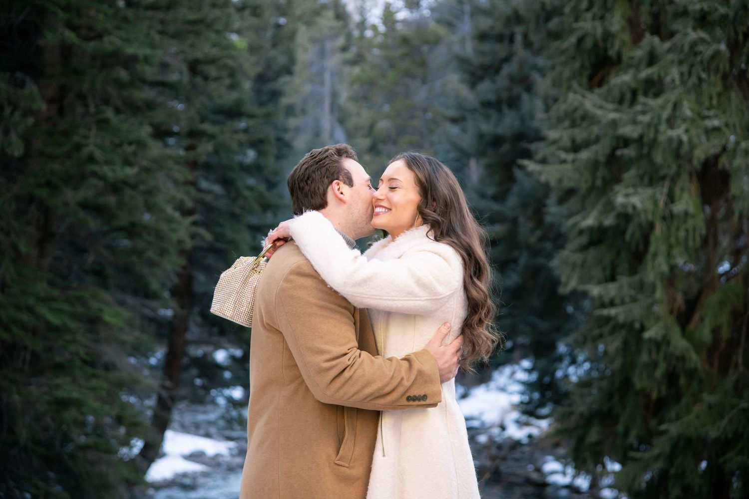 A couple shares a romantic kiss in a snowy winter forest setting with evergreen trees in the background for Vail Colorado engagement