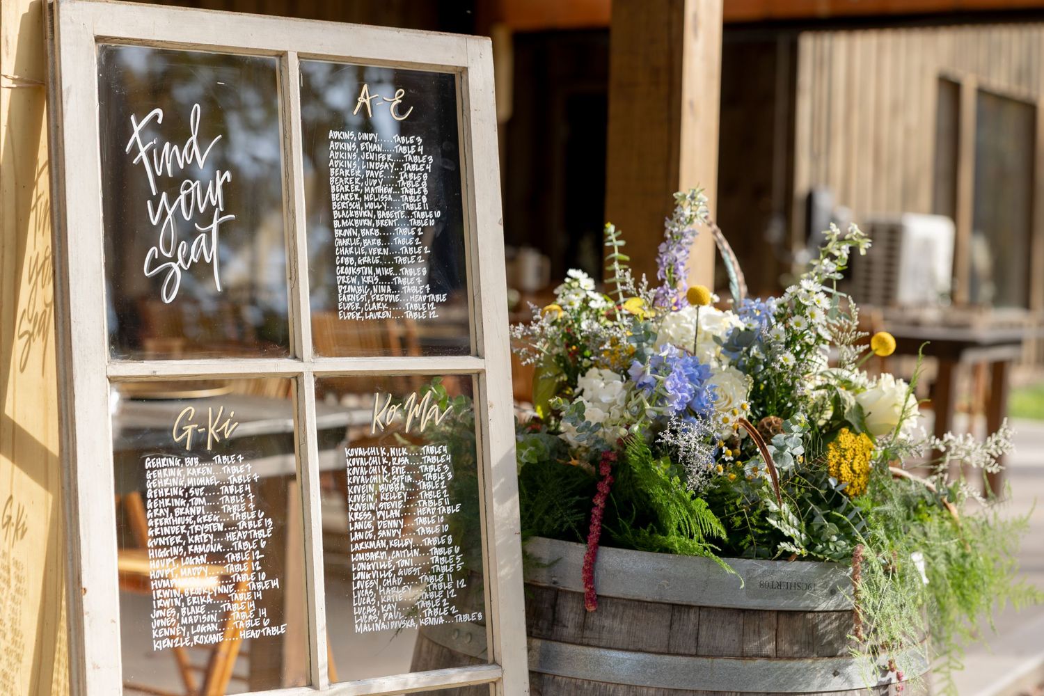 Wedding seating chart written in white text on glass windows with floral decorations and natural lighting.