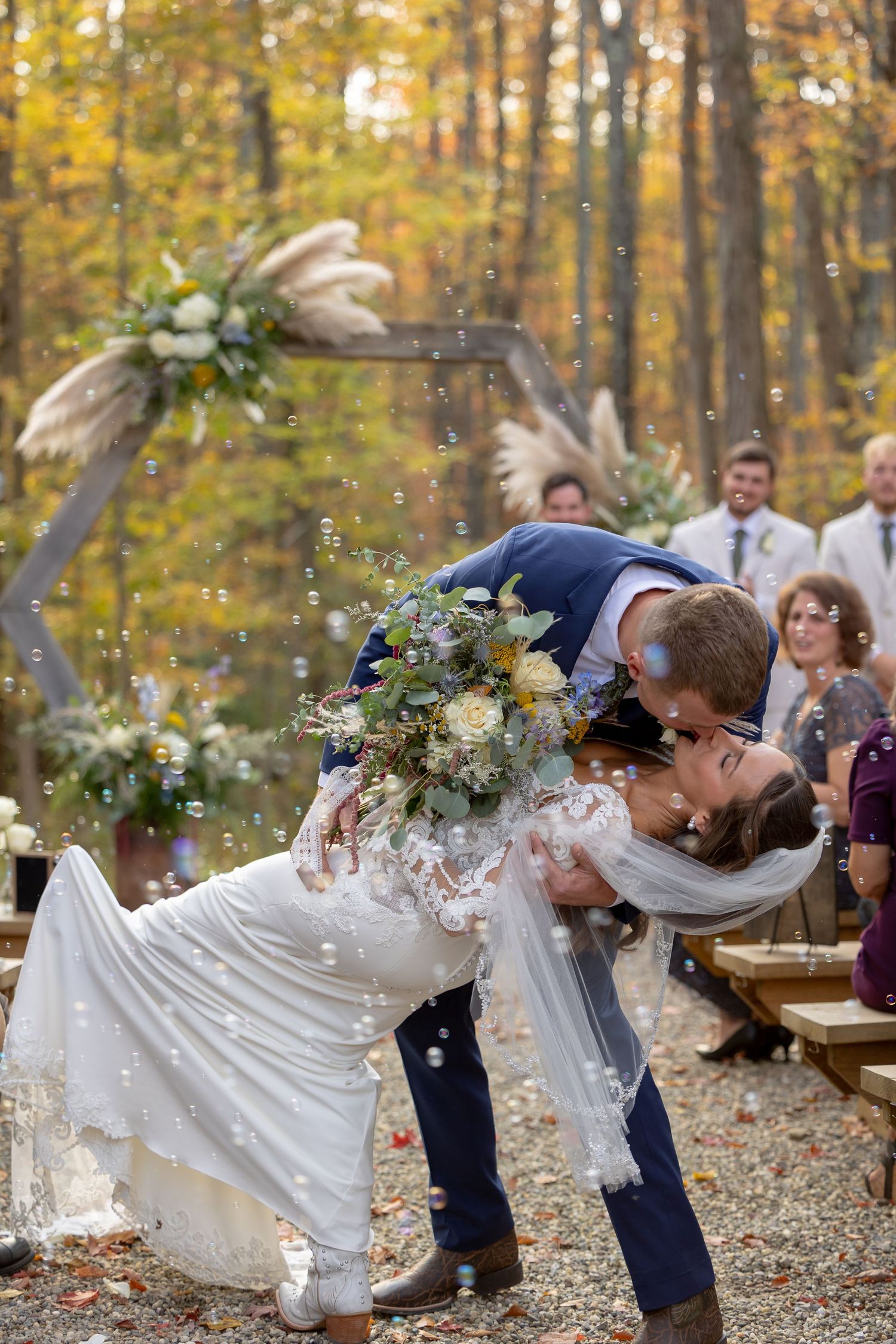 A wedding couple shares a romantic dip kiss in an autumn forest setting surrounded by golden leaves and rustic decor.