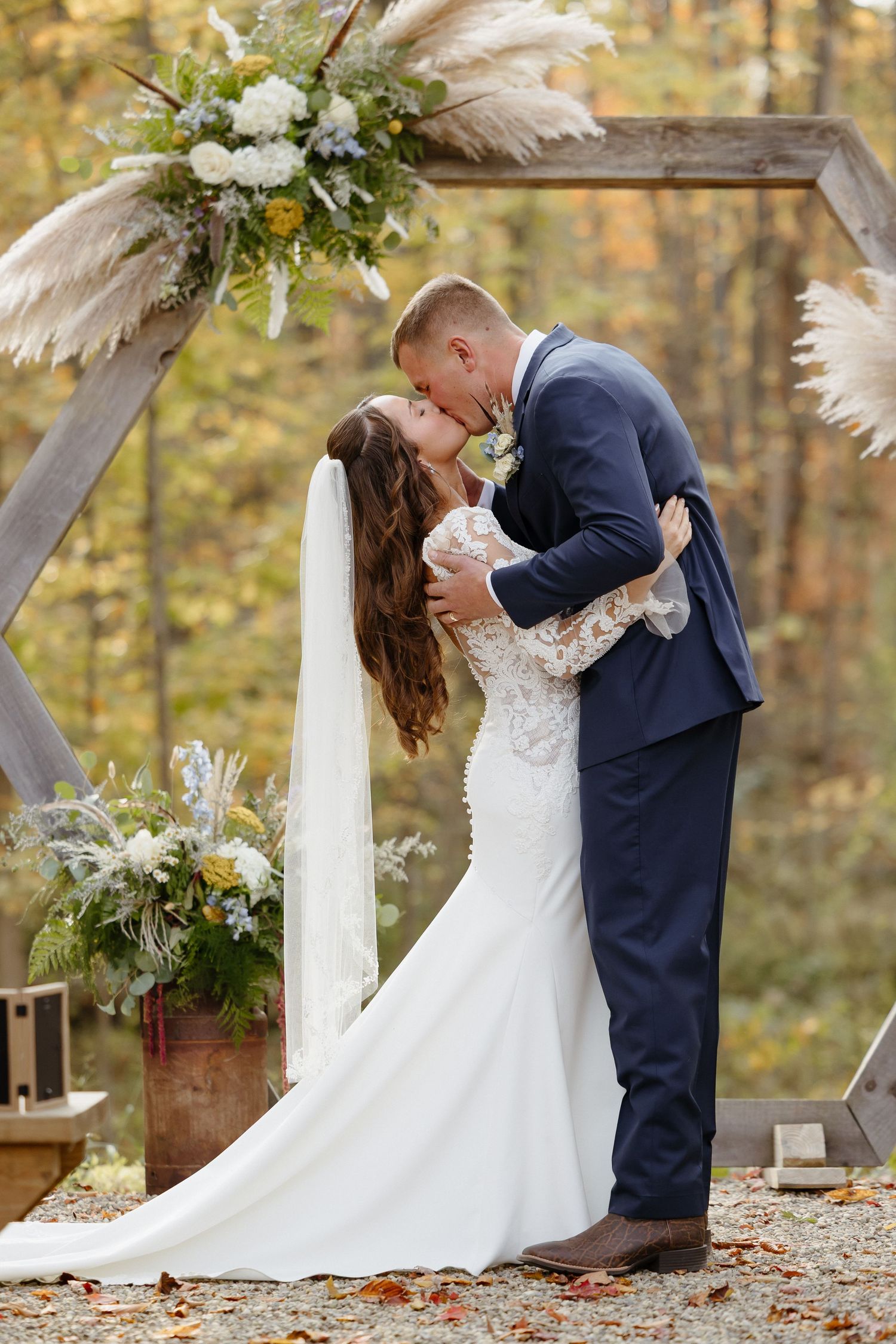 Romantic wedding couple shares a tender kiss under a floral arch during autumn outdoor ceremony.