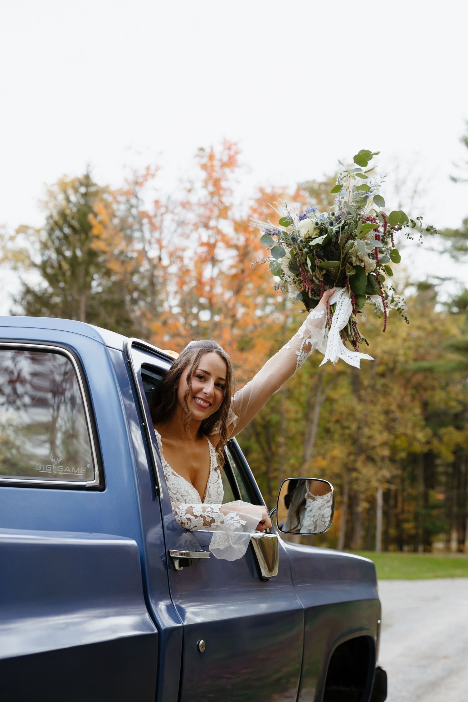 A joyful person in a wedding dress holds up a bouquet while leaning out of a vintage blue truck window on an autumn day.