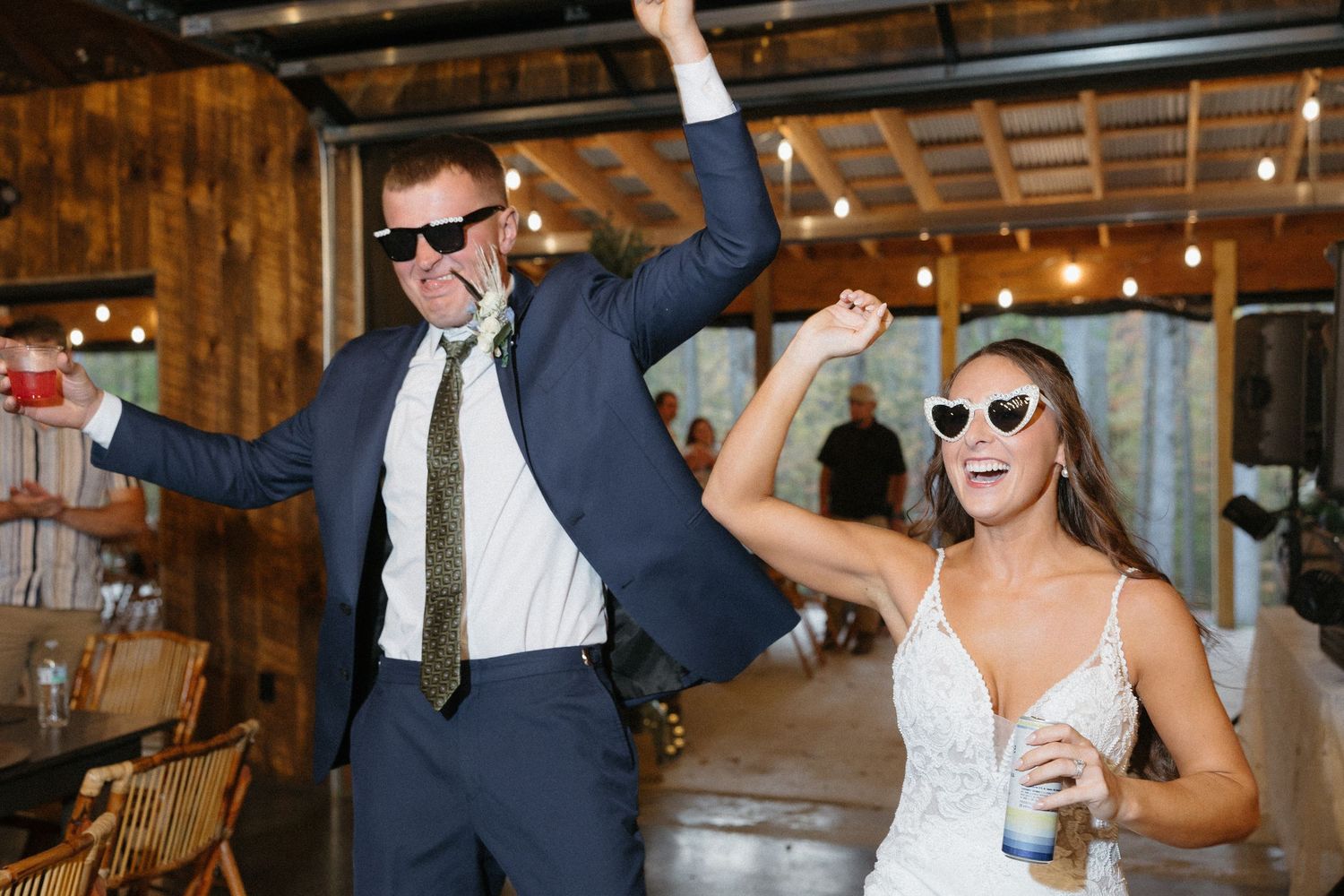 A couple wearing sunglasses dance joyfully at their rustic barn wedding reception with string lights overhead.